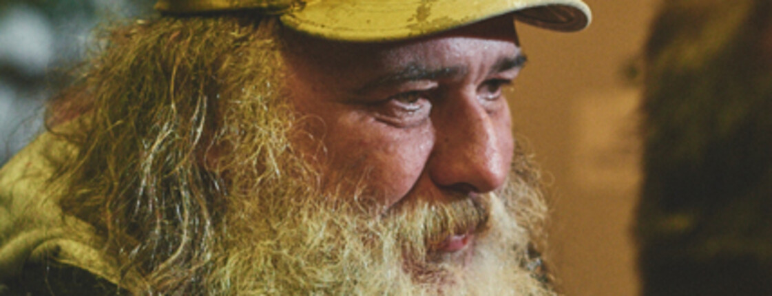 An elderly homeless man with a beard and a cap, engaged in conversation