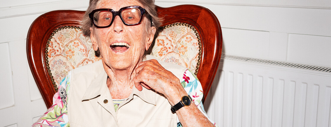 An elderly woman is sitting and smiling at the camera; she is wearing sunglasses.
